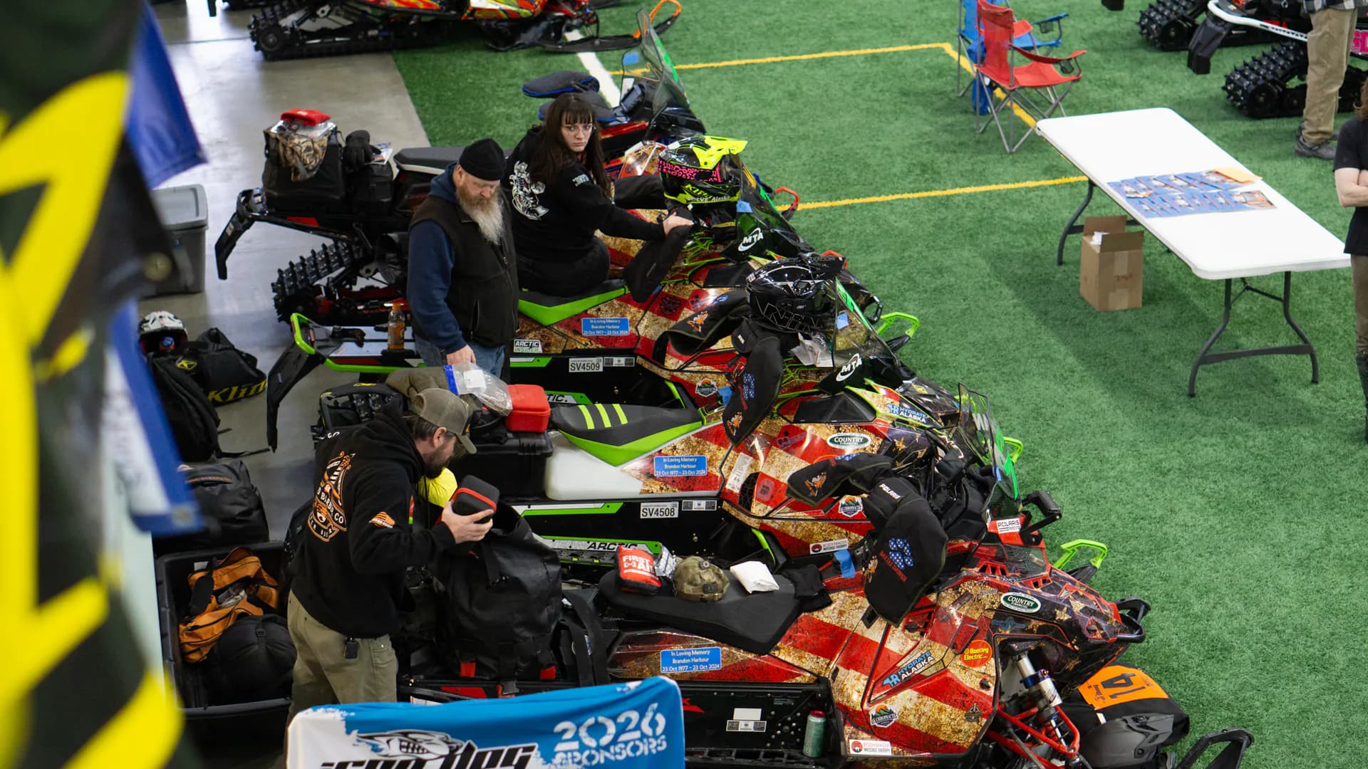 Wide view of the tech inspection hall with dozens of race sleds laid out across the turf.