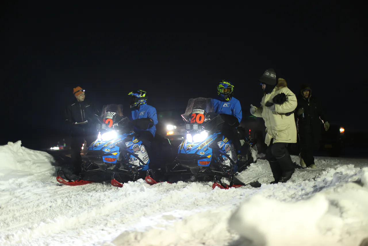 Teams pull in over a snow berm after dark, machine lights washing across the drift.