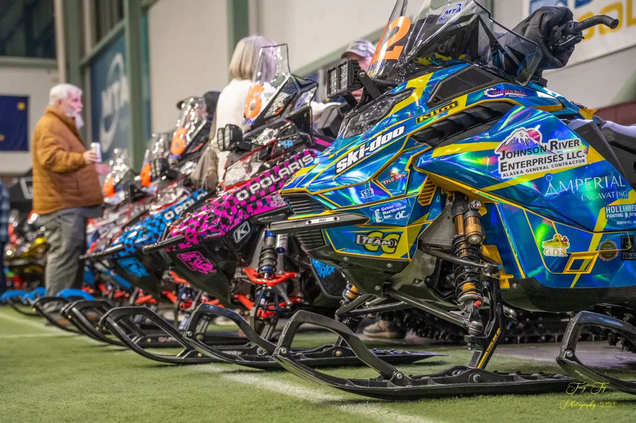 Race sleds on display indoors at the pre race expo with visitors walking the rows.
