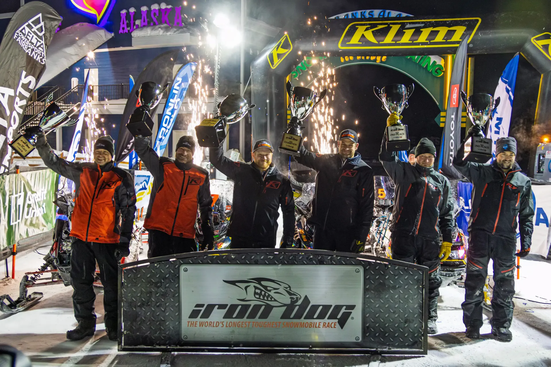 Podium finishers on the finish stage with arms raised as sparklers burn behind them.