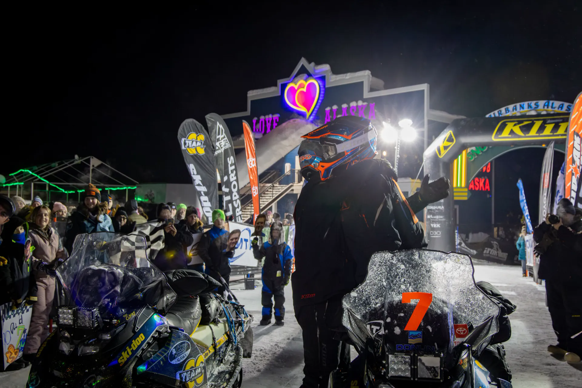 A team rolls into the Fairbanks finish after dark with the crowd and lights either side of the lane.