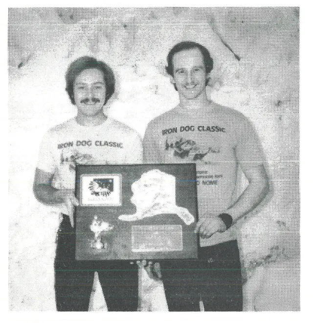 Two men in Iron Dog Classic t-shirts hold a plaque with a map of Alaska.