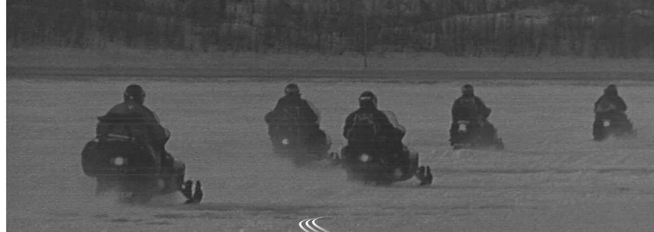 Five snowmachines run in a line across a frozen lake, timber on the far shore.