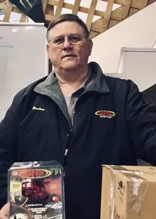 A man in a black team jacket stands indoors holding a boxed part, cartons stacked beside him.