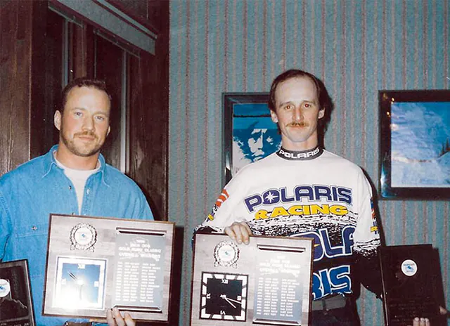 Two racers stand indoors holding engraved Iron Dog plaques, one of them in a Polaris Racing jersey.