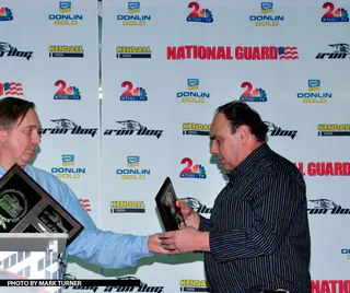 One man hands another an engraved plaque in front of an Iron Dog media wall at the awards ceremony.