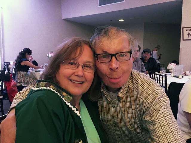 A couple lean their heads together for the camera at a banquet table, the room busy behind them.