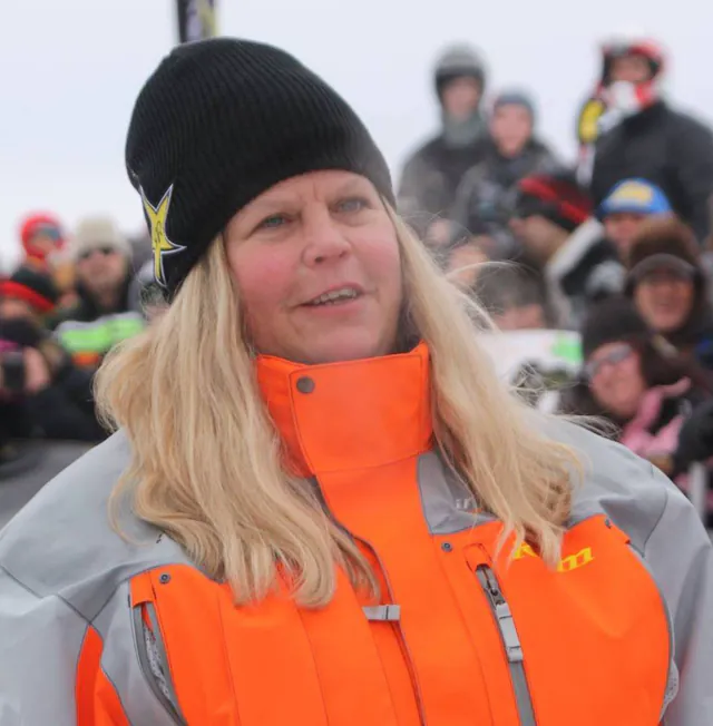 A rider in an orange and grey race jacket and a black beanie stands in front of a crowd of spectators on the snow.