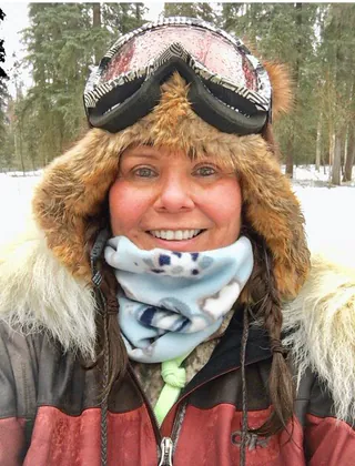 A woman in a fur ruffed hat with goggles pushed up and a printed neck warmer smiles in the snowy woods.