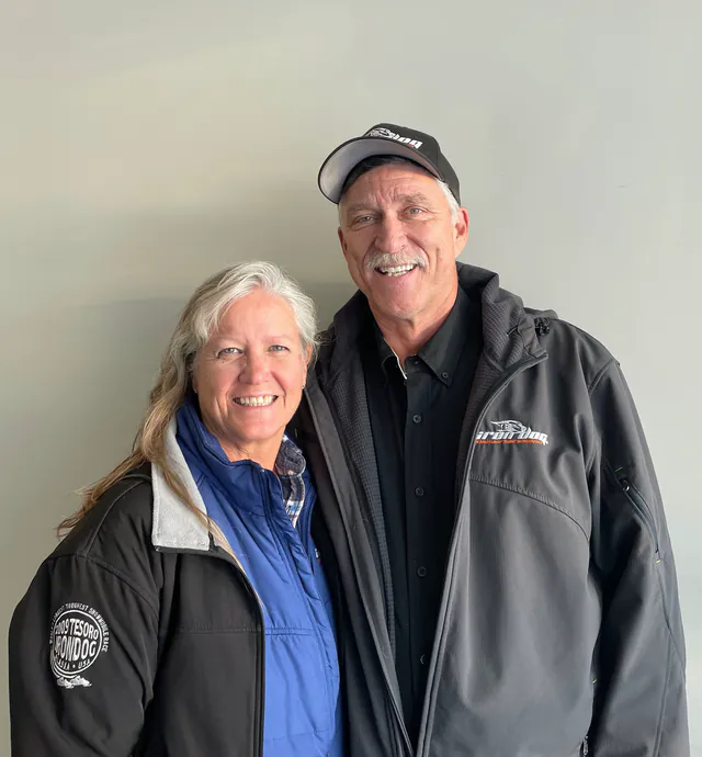 A couple in Iron Dog jackets stand together against a plain wall, the man in an Iron Dog cap.