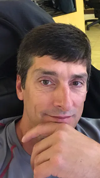 A man rests his chin on his hand and looks at the camera from a chair in an office.