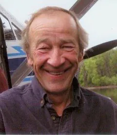 A grey haired man grins in front of a small plane, a river and spring trees behind him.