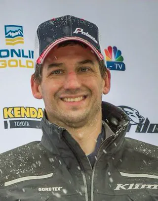 A racer in a Polaris cap and a snow flecked jacket stands in front of a race media wall.