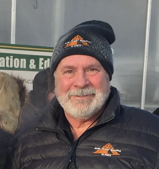 A bearded man in a black beanie and a down jacket smiles outdoors, a green sign and a white tent behind him.