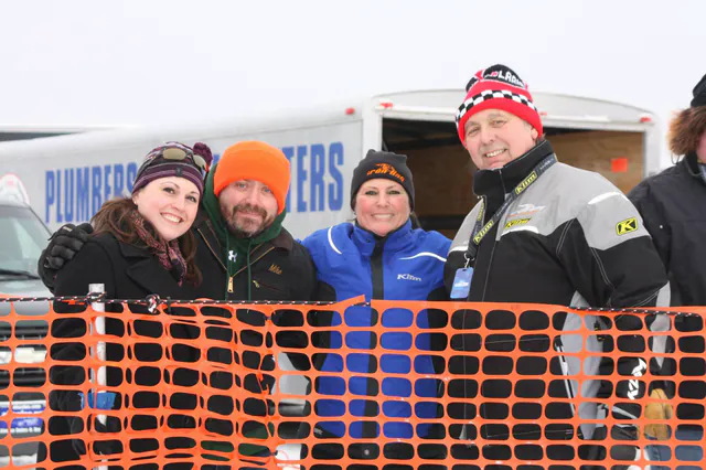 Four people in winter gear stand shoulder to shoulder behind an orange fence at the race, trailers parked behind them.