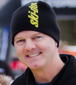 A man in a black Ski-Doo beanie smiles into the camera outdoors, a race machine out of focus behind him.