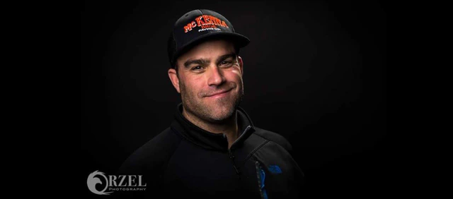 A racer in a black cap and a team jacket, lit against a black studio background.