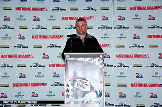 A man speaks from a lectern in front of an Iron Dog media wall, a large photograph of a race machine mounted on the front of the lectern.