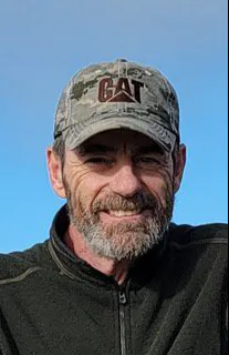 A man in a camouflage cap smiles against a clear blue sky.