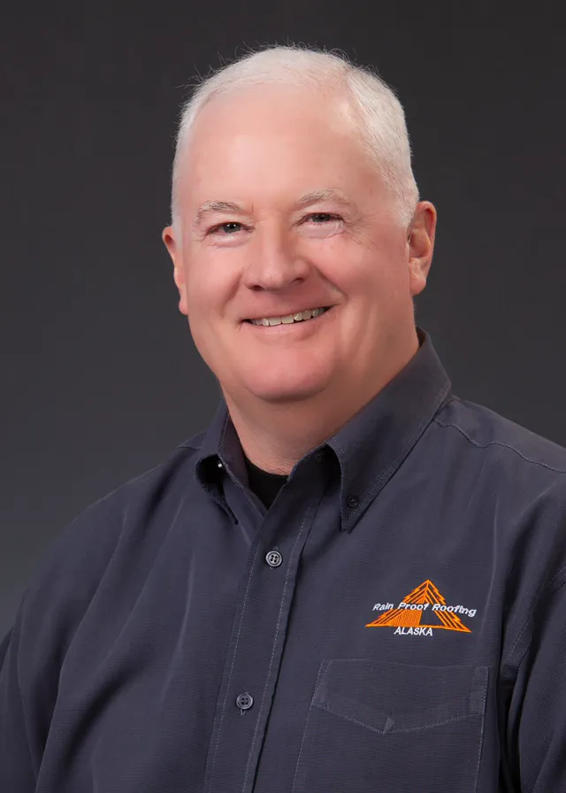 A studio portrait of a white haired man in a grey work shirt against a dark grey background.
