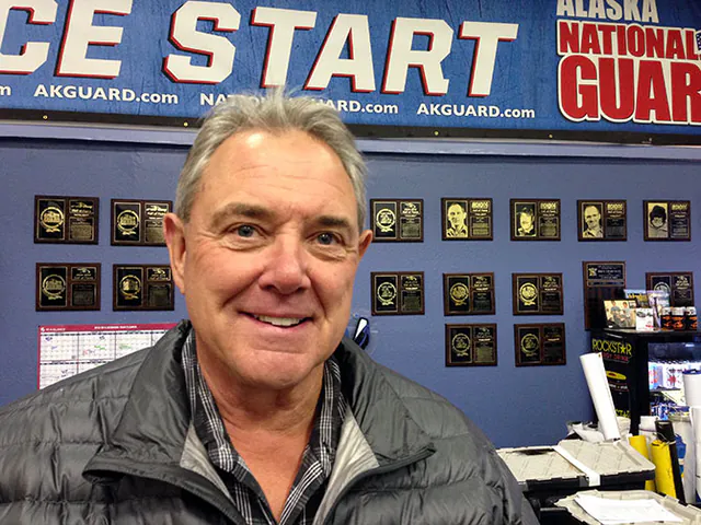 A man stands in front of a wall of engraved plaques inside a shop, an Alaska National Guard race sign above him.