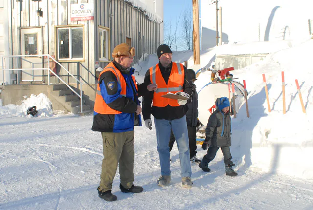 Two men in orange hi-vis vests stand talking on packed snow outside the Crowley building at Galena, one holding a clipboard, a fuel tanker and a snow berm behind them and a child in a blue hat walking past.