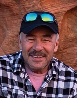 A man in a plaid shirt and a cap with sunglasses pushed onto the brim smiles in front of a red rock wall.