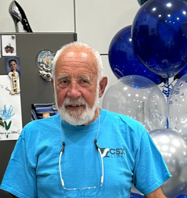A white bearded man in a bright blue shirt stands beside a bunch of blue and silver balloons in an office.