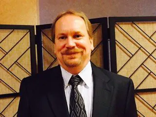 A man in a dark suit and a patterned tie stands in front of a woven screen indoors.