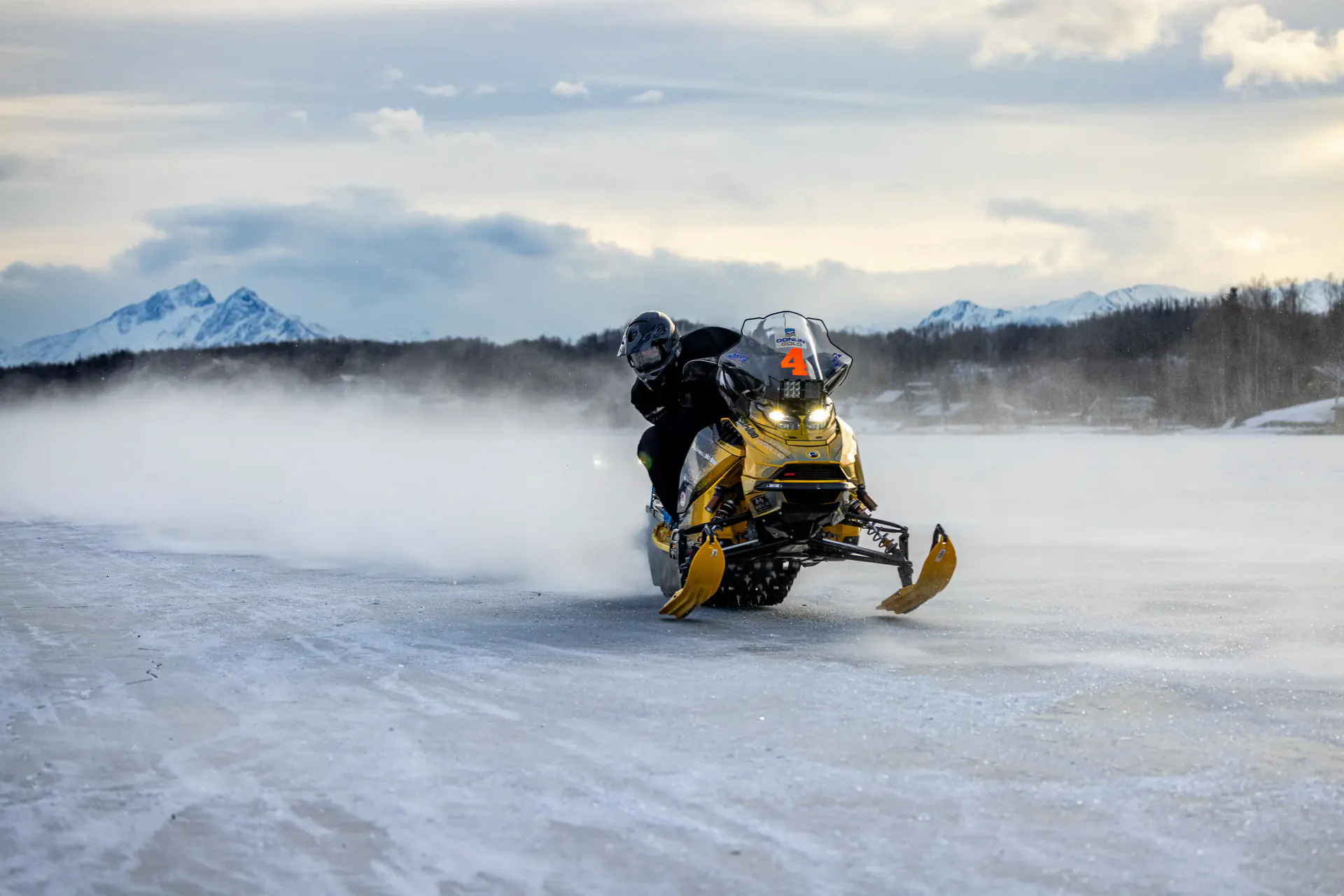 A lone racer throws up a wall of snow crossing a frozen lake at dusk, mountains behind.
