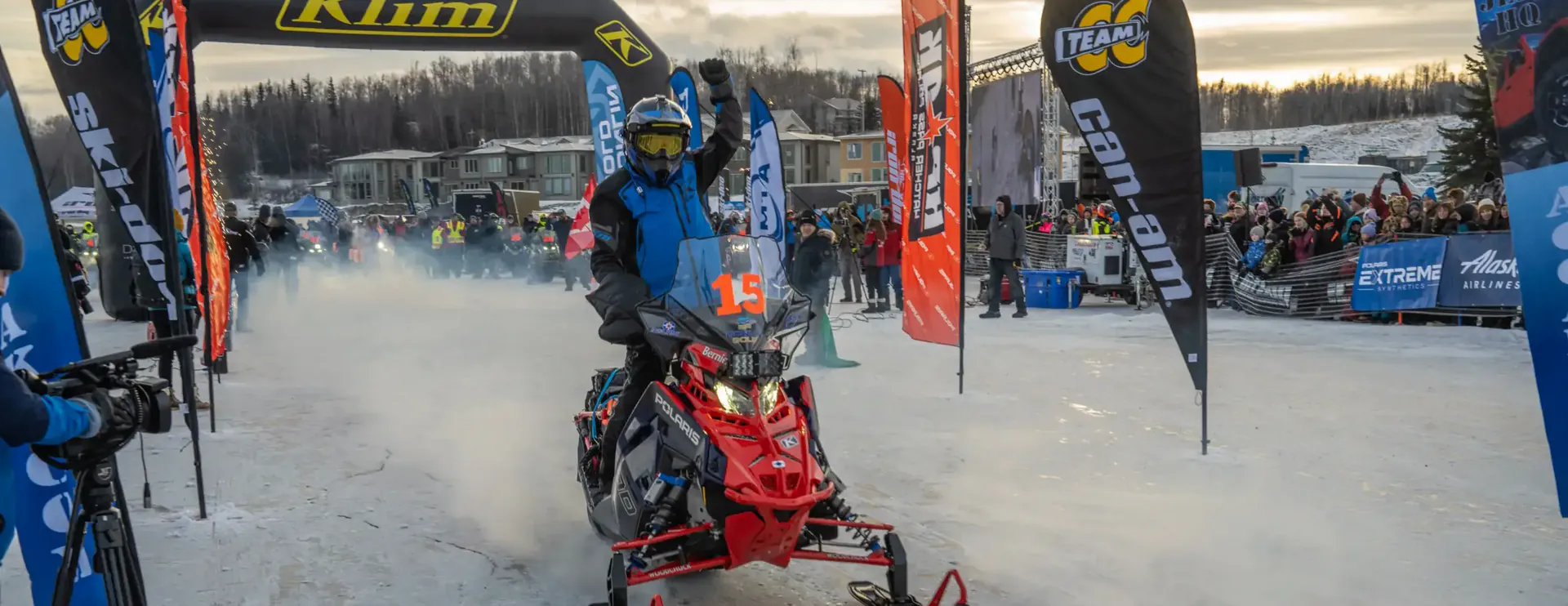 Ultra wide view along the Big Lake start chute as a racer stands on the running boards with one arm raised.