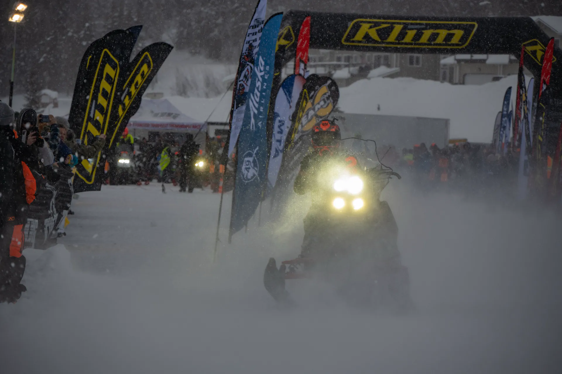 A team pulls away from the Big Lake start line into heavy falling snow, machine lights cutting through the flag line.