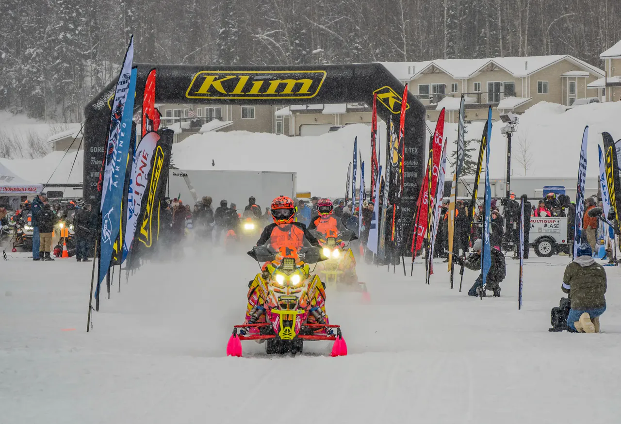 A single sled accelerates out of the start chute in a blizzard with spectators and flags either side.