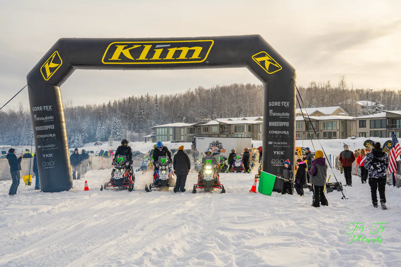 Expedition Class sleds gathered under the start arch at Big Lake on a clear afternoon.