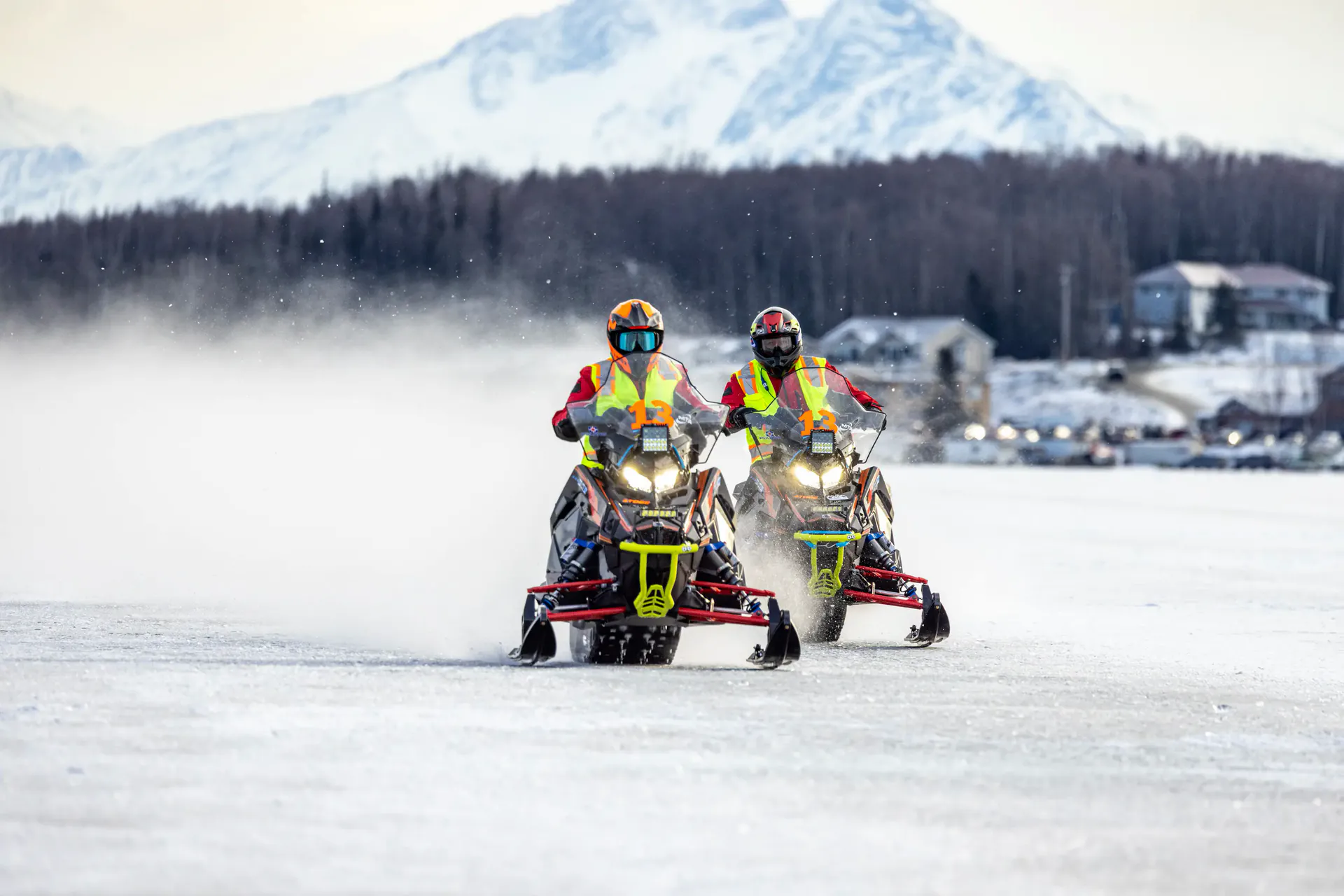 Two Pro Class race sleds running side by side across open lake ice, snow spray trailing behind them and mountains on the horizon.