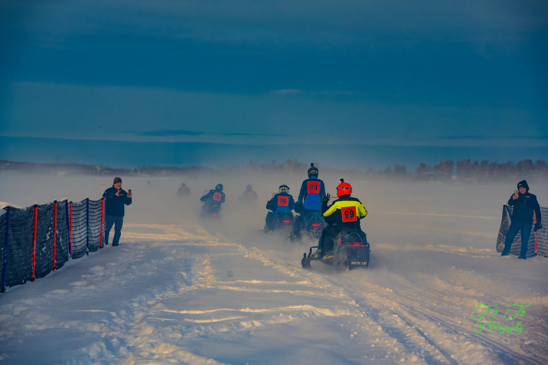 Riders disappear into ice fog out on the lake while spectators watch from the fence line.