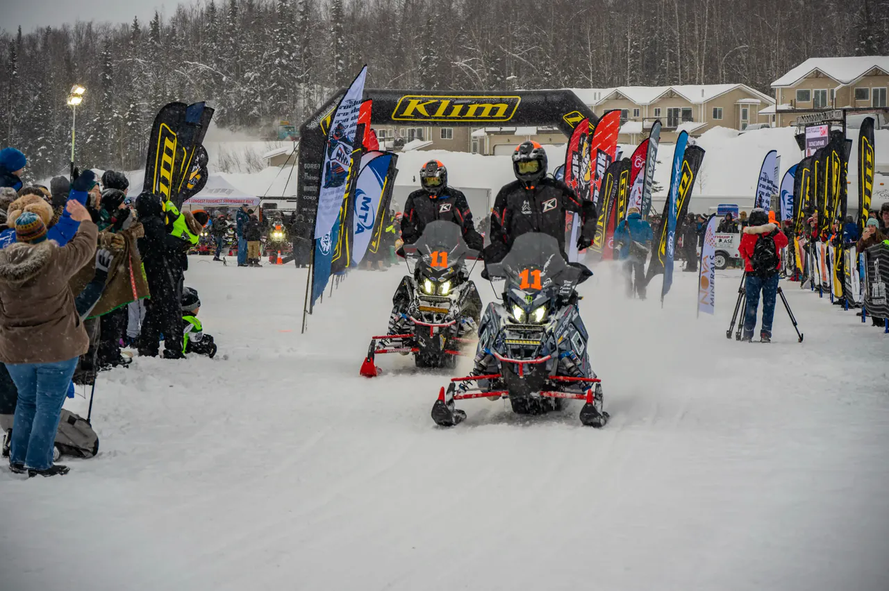 Two race sleds leave the Big Lake start line together as photographers kneel in the snow.