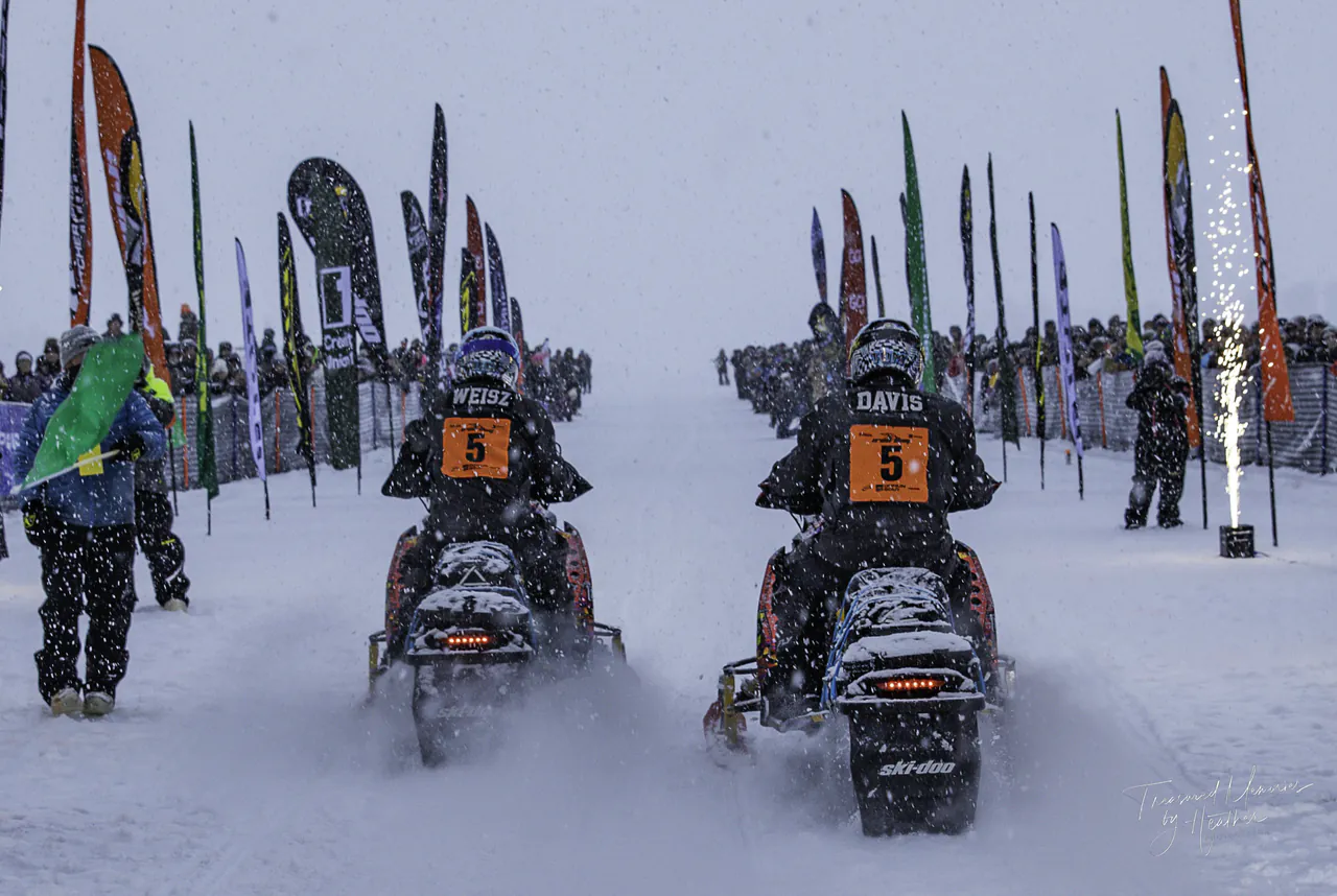 A pair of race sleds staged side by side in falling snow with feather flags lining the chute.