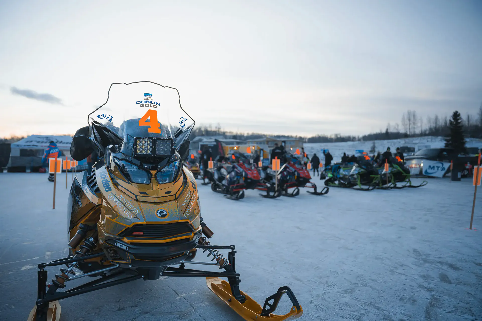A race sled staged in the start chute at Big Lake with crews and spectators along the fence.