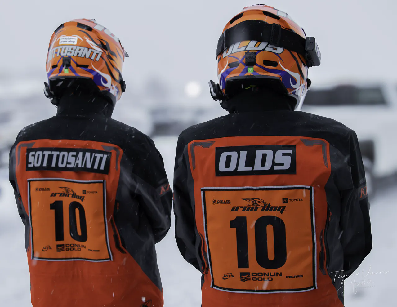 Two teammates seen from behind in matching orange race bibs numbered 10, helmets on, waiting to start.