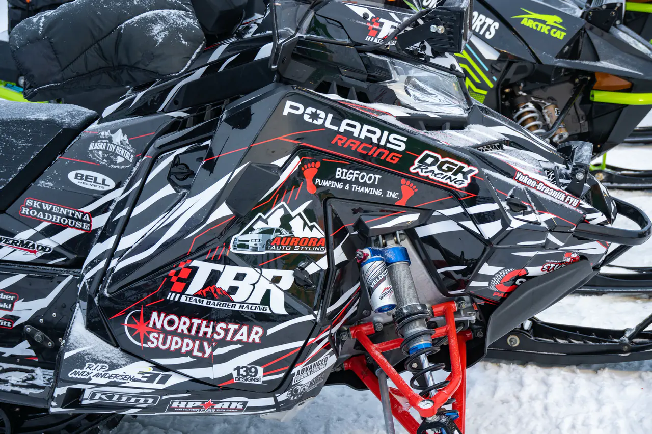 Close view of a race sled cowl and front suspension under fresh snow, supporter decals across the panel.