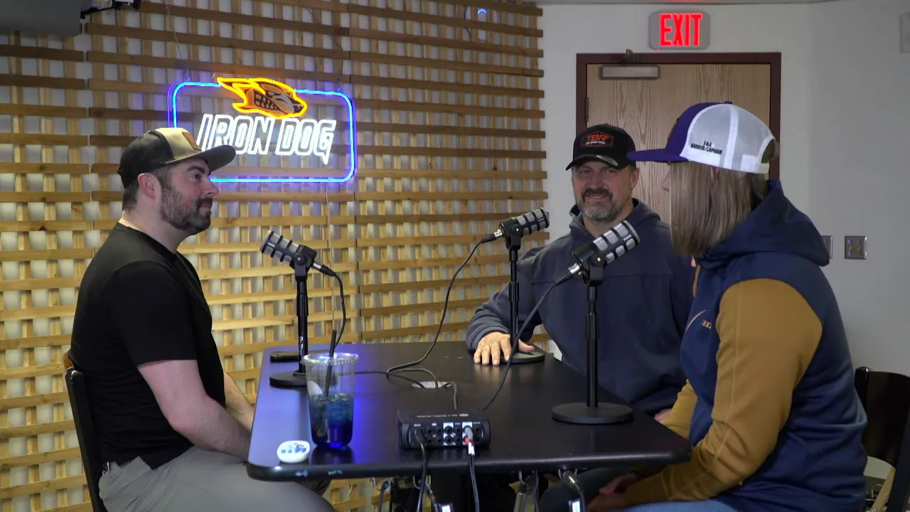 Three people at microphones around a table in the recording room, a lit Iron Dog sign on the lattice wall behind them.