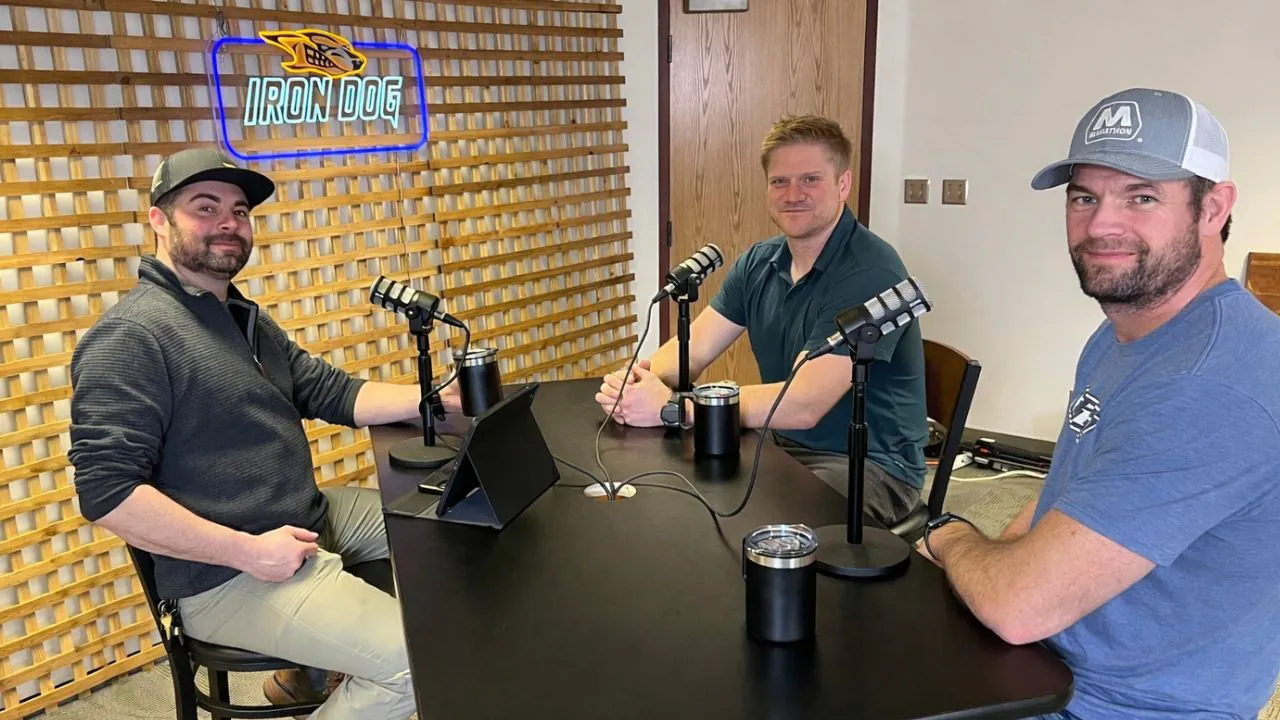 Three men seated around the microphones turn to the camera, the lit Iron Dog sign on the wall beside them.