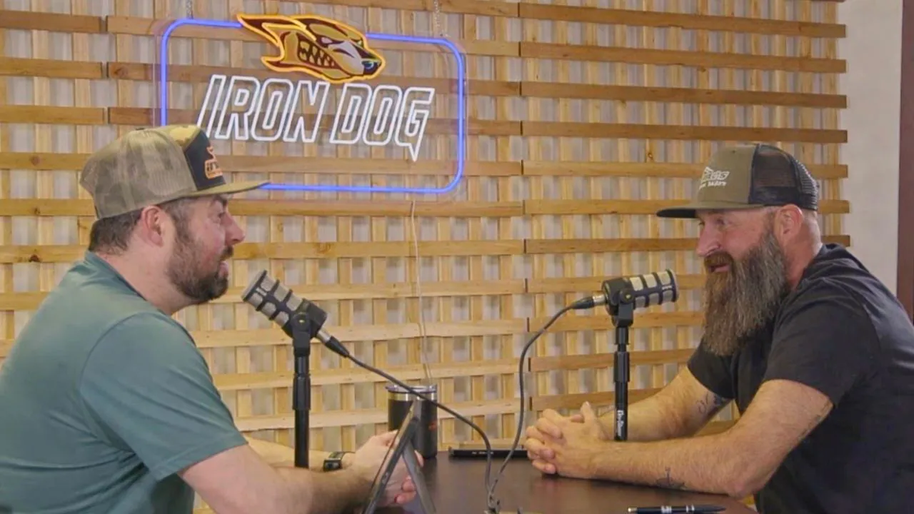 Two men face each other across the microphones under the lit Iron Dog sign, one of them with a long beard.