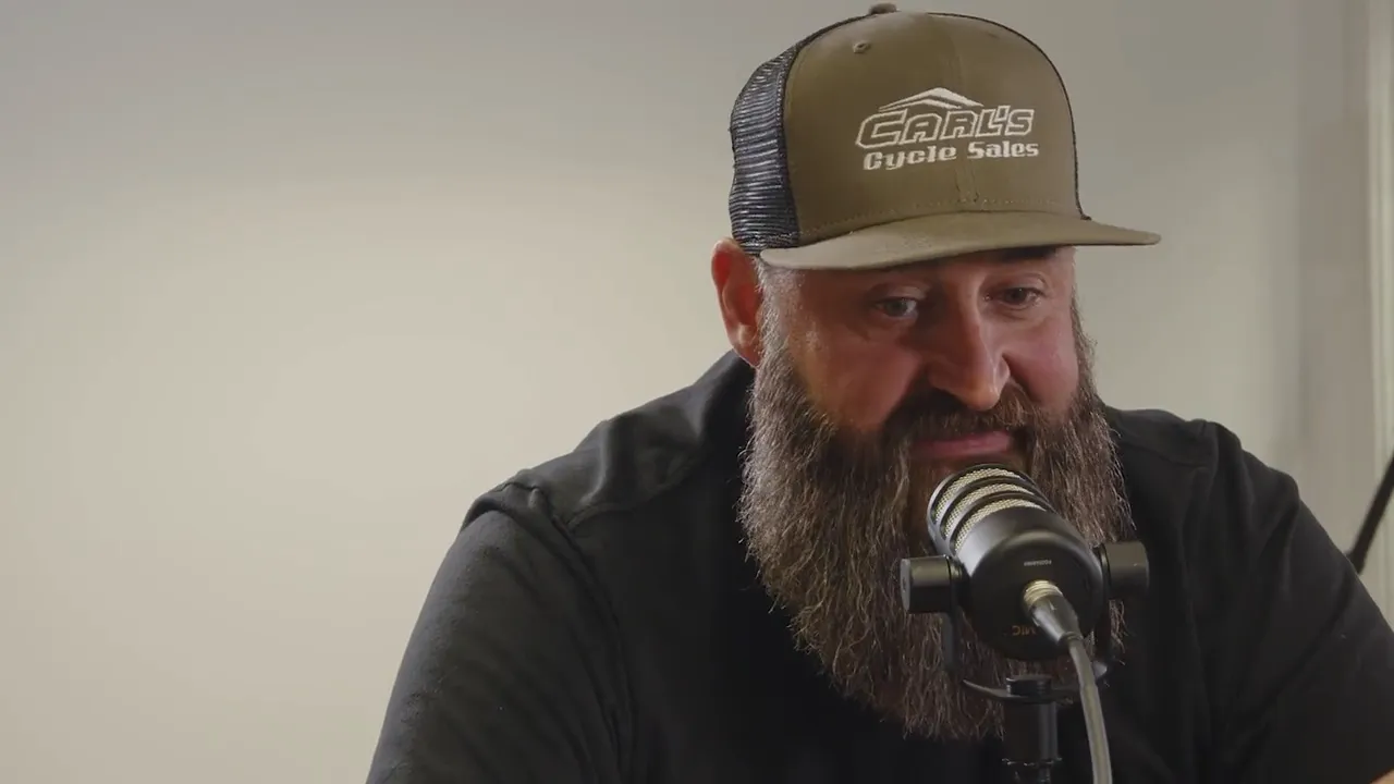 A bearded racer in a Carl's Cycle Sales cap speaks into a studio microphone against a pale wall.