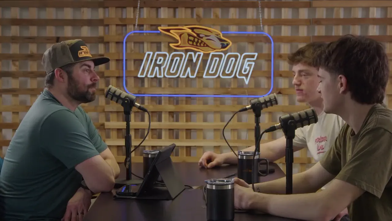 A wide view of the recording room: a lit Iron Dog sign on a wooden lattice wall, three microphones on the table and a host facing two guests.