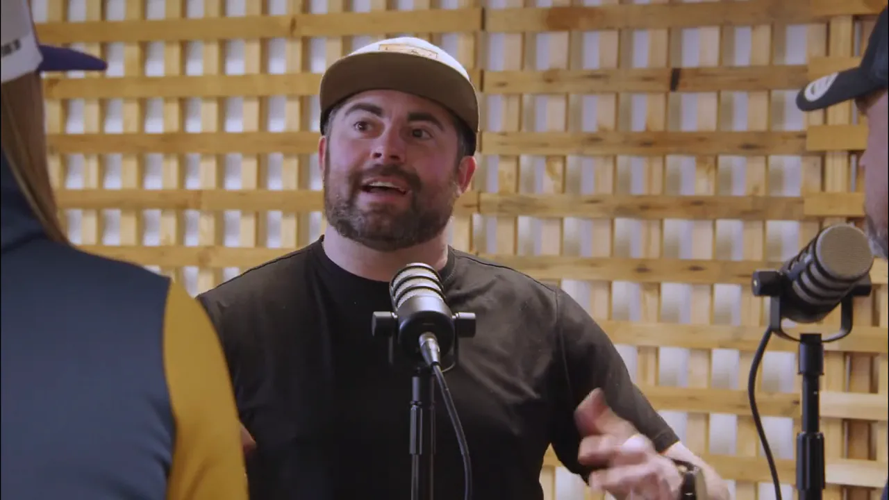 A guest in a cap talks across the microphone with one hand raised, the wooden lattice wall of the recording room behind him.