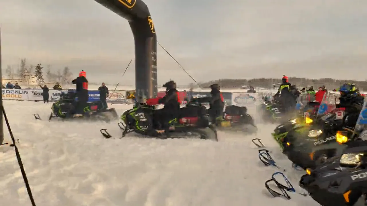Riders in orange climb a snow bank through the trees, a machine kicks ice across a frozen lake, then the start chute under a black arch and a sled on a red stand in the shop.