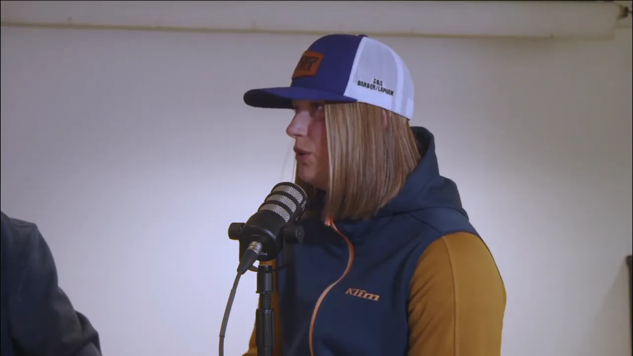 A racer in a team cap and a navy and gold jacket speaks into the microphone in profile against a plain wall.