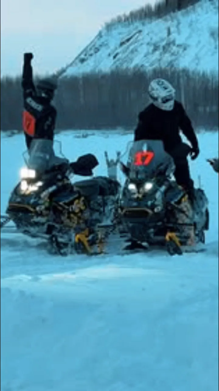 Shot from a moving machine: a night trail, open lake ice at sunset, then two riders on bib 17 standing on their sleds with a fist in the air, a snow bluff and black timber behind them.